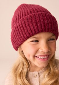 Dunkelroter gestrickter Beanie mit vertikaler Rippstruktur. Abgerundete Oberseite und enger Sitz. Wird von einem Mädchen mit langen blonden Haaren getragen.