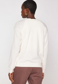 Pull crème à manches longues, avec des poignets côtelés et une coupe décontractée. Porté avec un pantalon marron, mettant en avant une texture douce et un design simple.