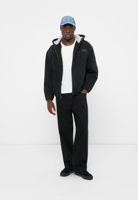 Svart fleece-fôret jakke med hette, hvit t-skjorte, svarte bukser og svarte sneakers. Blå caps med logo tilfører fargekontrast.