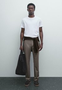 Jonge man in een wit T-shirt, beige broek, met een bruine trui om zijn taille geknoopt, houdt een grote zwarte totebag vast, staande tegen een effen lichte muur.