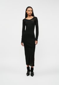 Dopasowana czarna sukienka z długimi przezroczystymi rękawami i okrągłym dekoltem. Gładka tekstura, długość do połowy łydki, rozcięcia po bokach, noszona z botkami na obcasie.