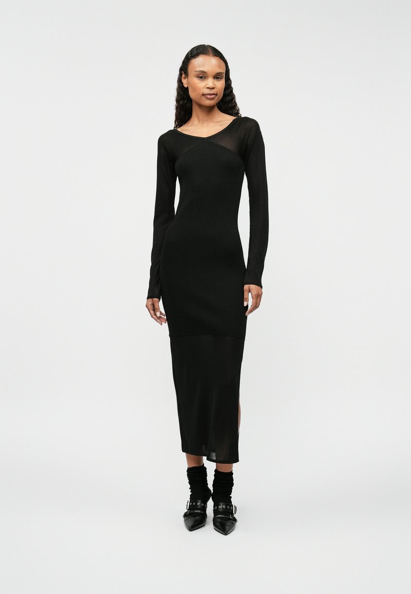 Dopasowana czarna sukienka z długimi przezroczystymi rękawami i okrągłym dekoltem. Gładka tekstura, długość do połowy łydki, rozcięcia po bokach, noszona z botkami na obcasie.