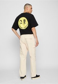 Czarny T-shirt z krótkim rękawem z dużym żółtym nadrukiem na plecach, zestawiony z beżowymi spodenkami typu cargo i czarnymi trampkami.