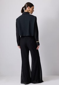 Afskåret sort jakke med høj krave over et sort jumpsuit med brede ben. Gennemsigtig stofdetaljer og guldfarvede metaldele på ærmerne.
