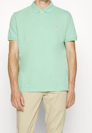 Mann trägt ein hellgrünes Poloshirt mit kurzen Ärmeln und kleinem orangefarbenem Logo auf der Brust sowie beige Hose, steht vor weißem Hintergrund.