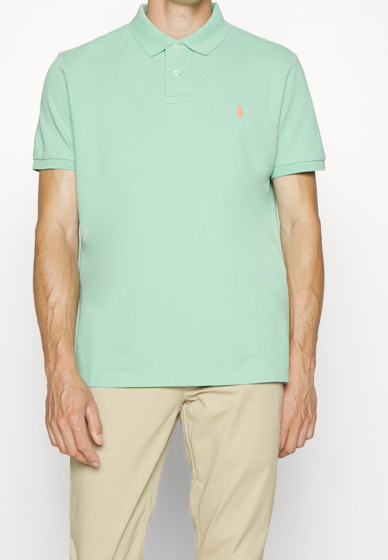 Homme portant un polo vert clair à manches courtes avec un petit logo orange sur la poitrine et un pantalon beige, debout sur un fond blanc.