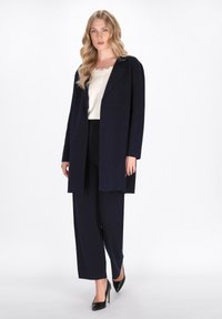 Cappotto lungo in misto lana blu navy con una vestibilità rilassata, abbinato a un top crema con bordi in pizzo e pantaloni a gamba larga coordinati; tacchi alti neri completano il look.