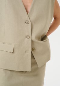 Gaišs khaki bezpiedurkņu žaketē ar dziļu V-veida kakla izgriezumu, trim pogām un sānu kabatu, ar vieglu tekstūru.