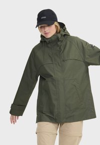 Veste imperméable vert olive avec une capuche, des cordons de serrage réglables, des poches avant et des fermetures à pression. Associée à un pantalon cargo beige.