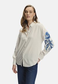 Camisa blanca de botones con bordado floral azul en la manga izquierda, con un cuello clásico y mangas largas, hecha de tela ligera.