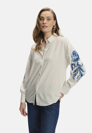 Camisa blanca de botones con bordado floral azul en la manga izquierda, con un cuello clásico y mangas largas, hecha de tela ligera.