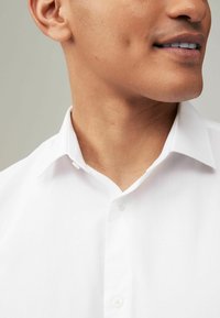 Weißes Hemd mit klassischem Kragen, glatter Textur und einem einzelnen Knopf am Ausschnitt sichtbar. Subtile glänzende Oberfläche des Stoffes.