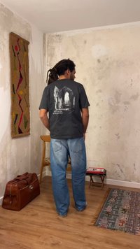 Άνδρας με dreadlocks, που φοράει μαύρο μπλουζάκι με γραφικό σχέδιο και μπλε τζιν, στέκεται σε δωμάτιο με ξύλινη καρέκλα, δερμάτινη τσάντα, μοτίβο σε χαλί και υφασμένους τοίχους.