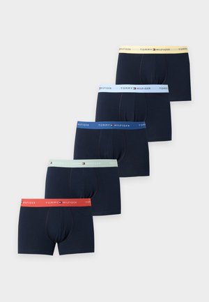 Homme noir debout aux cheveux courts et bouclés, portant un boxer sombre avec une ceinture claire sur un fond clair uni.