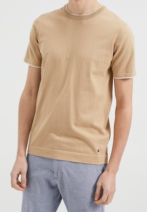 Homme portant une chemise tricotée beige à manches courtes avec des bordures blanches sur les manches et un pantalon gris clair, devant un fond uni.