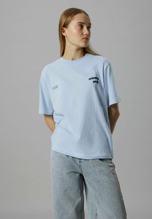 Ung kvinde med langt blondt hår, iført en lyseblå oversized T-shirt og lyse jeans, der kigger til siden mod en grå baggrund.