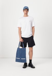 Denim tote bag with two handles, featuring white "SAMSØE" text, worn with a white t-shirt, black shorts, and black shoes.