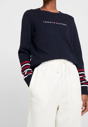 Osoba nosząca granatowy sweter Tommy Hilfiger z mankietami w czerwone, białe i granatowe paski oraz białe spodnie ze sznurkiem.
