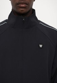Marinblå zip-jacka med hög krage, texturerad tyg och subtila ljusblå sidostråk. Har en cirkulär logotyp på bröstet.