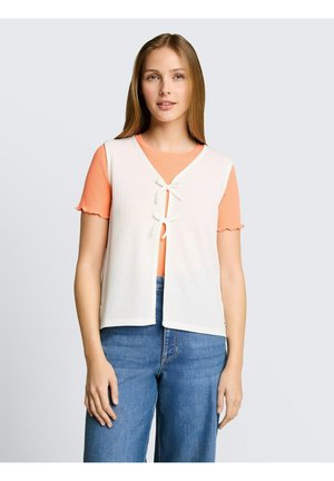 Junge Frau mit glatten hellbraunen Haaren, die eine weiße gestrickte Weste mit Schleifen über einem orangen kurzärmligen Oberteil und blauen Jeans trägt.