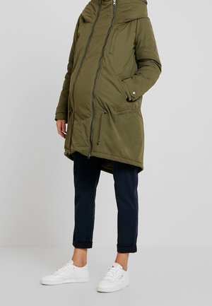 Parka acolchada verde oliva con capucha, dos bolsillos frontales, cierre de doble cremallera y dobladillo ajustable con cordón. Combinada con pantalones negros con puños enrollados y zapatillas blancas.