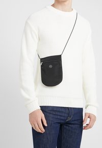 Svart nylon crossbody-väska med dragkedjestängning och logodetalj, bärs över en krämfärgad stickad tröja och blåjeans. Enkel, kompakt design.