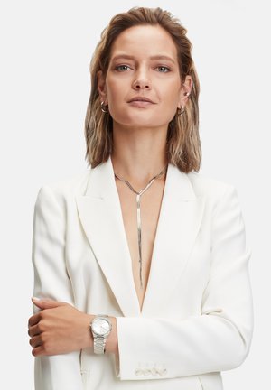 Zilveren ketting met een slank, langwerpig ontwerp, gedragen met een witte blazer. Het model draagt ook een zilveren horloge met een klassieke wijzerplaat.