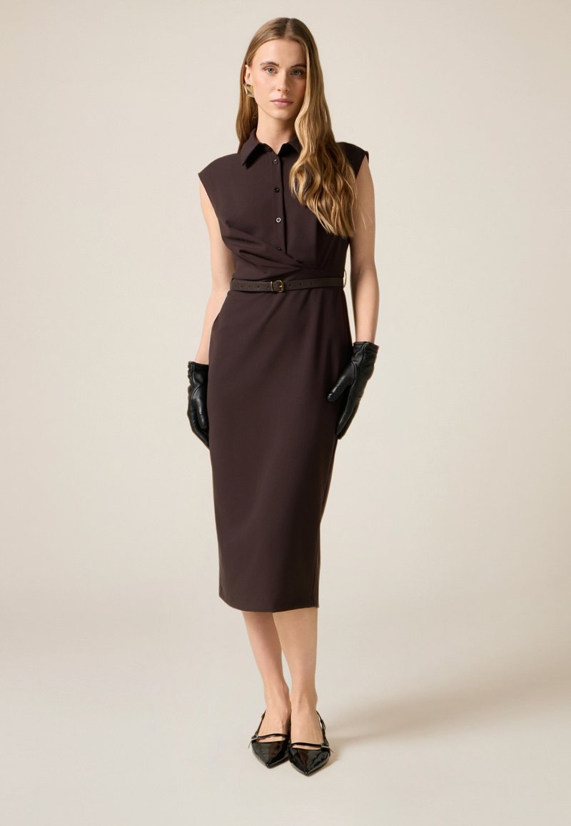 Brun, skræddersyet kjole med krave, korte ærmer og bælte i taljen. Glat stof, knælængde, båret med sorte handsker og spidse sko.