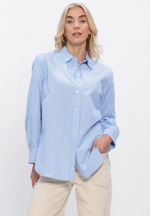 Lyshåret kvinde iført lyseblå, stribet skjorte med knapper og beige bukser, poserer med den ene hånd i lommen mod en hvid baggrund.