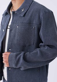 Giacca blu navy testurizzata con vestibilità squadrata, caratterizzata da collo, due tasche frontali, dettagli in metallo argentato e chiusure con bottoni.