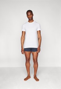 T-shirt en coton blanc à manches courtes, associé à un short de boxeur bleu marine avec un motif subtil. Le mannequin se tient pieds nus contre un fond neutre.