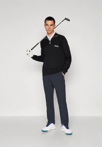 Μαύρο πλεκτό πουλόβερ με λευκό γιακά και λογότυπο "BOSS", συνδυασμένο με μπλε σκούρο παντελόνι και λευκά παπούτσια γκολφ, κρατώντας μια ράβδο γκολφ, στέκεται μπροστά σε απλό φόντο.