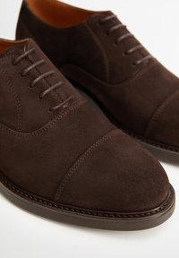 Chaussures habillées en daim marron foncé avec lacets, dotées de détails cousus et de semelles empilées basses sur un fond blanc.