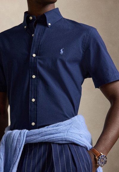 Muž nosi tmavomodrú krátku košeľu s gombíkmi a logom, pásikavé nohavice, svetlomodrý sveter zviazaný v páse a hnedé kožené hodinky na zápästí.