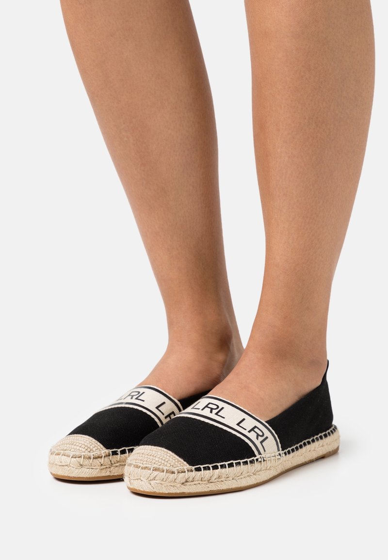 Jambes portant des espadrilles noires avec des semelles en corde beige et des bandes beiges brandées LRL sur le dessus, debout sur une surface blanche.