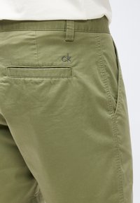 Κοντινή λήψη στην τσέπη πράσινων σκούρων παντελονιών με θηλιά για ζώνη, πλαϊνή τσέπη στη ραφή και μικρό κεντητό λογότυπο "ck" πάνω από την πίσω τσέπη.