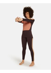Vrouw met krullend haar die haar armen naar voren strekt, gekleed in een donkerbruine en perzikkleurige lange mouwen top en bijpassende legging met een zijstrepen, blote voeten.