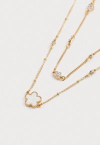 Zlatý náhrdelník so zlatým tónom s dvoma prameňmi, ktorý má prívesok v tvare kvetiny a priehľadné kryštály na článkoch reťazca.