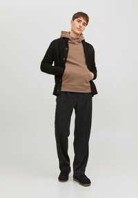 Cardigan nero lavorato a maglia con un motivo testurizzato, indossato sopra un hoodie marrone. Pantaloni neri slim-fit e scarpe slip-on nere completano il look.