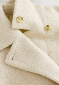 Cream-colored coat with a textured, fuzzy fabric. Features a notched collar and two rounded buttons with a subtle pattern.
