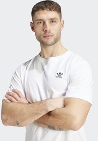 Bílé bavlněné tričko s malým černým logem Adidas na levé straně hrudi, kulatým výstřihem a krátkými rukávy. Hladký materiál a volný střih.