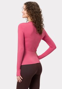 Langärmliges, tailliertes Shirt in leuchtendem Pink, aus dehnbarem Stoff gefertigt. Hat einen gewellten Saum und ein nahtloses Design auf der Rückseite. Kombiniert mit braunen Leggings.