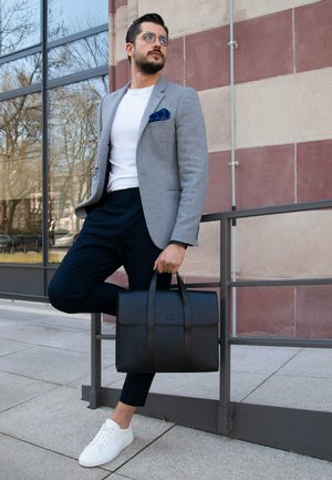 Gray houndstooth blazer, white t-shirt, navy trousers, white sneakers, and a black leather briefcase with side handles. Model leans against railing.
