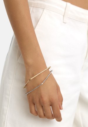 Dwie cienkie bransoletki na nadgarstku: jedna złota, a druga srebrna. Obie mają gładki, elegancki design oraz delikatny, minimalistyczny wygląd.