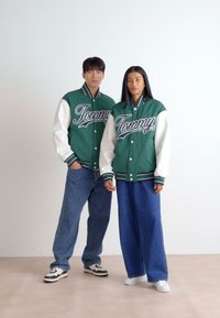 Jaquetas varsity verdes e brancas com o logótipo "Tommy", combinadas com jeans de corte largo. As jaquetas têm punhos de ribana listados.