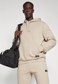 Sweatshirt bege com bolso canguru e cordão ajustável, combinado com calças bege a condizer. Mala de desporto preta com detalhes em rede.