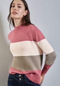 Randig tröja i rosa, krämfärgad och grön med ribbad hög krage och muddar, gjord av mjukt tyg; bärs med mörka jeans.