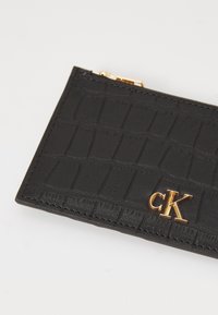 Portafoglio in pelle nera con una texture a coccodrillo, logo dorato "CK" e chiusura con zip; presenta cuciture ordinate e un design piatto.