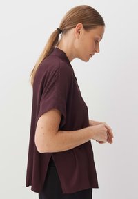 Burgundy short-sleeve blouse with a collar, loose fit, and back vents, made from soft fabric, featuring a smooth texture and clean lines.
