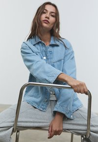 Svetlo modra jeans jakna z sproščenim krojem, bakrenim okovjem in dvema žepoma na prsih. Model sedi sproščeno na kromirani stoli. Sive trenirke.